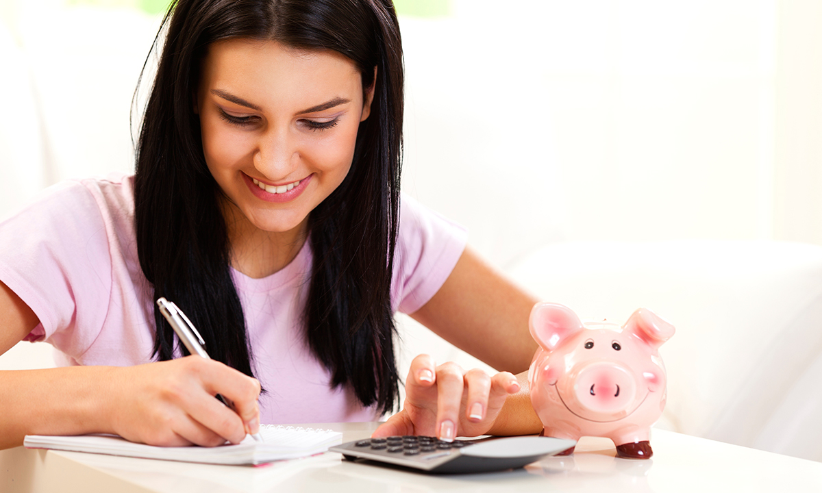 Woman writing in a notebook, with a calculator and piggy bank next to her