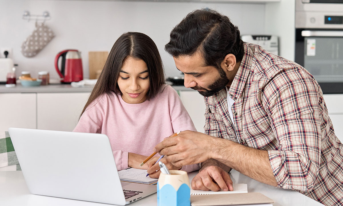 A father sits at a table with his daughter helping with her homework